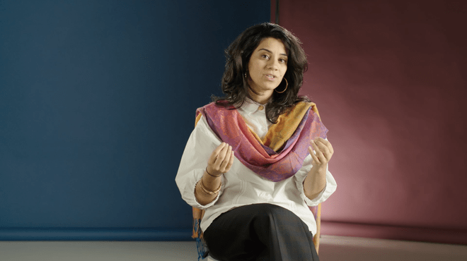 Screenshot of a moving image showing a woman talking towards the camera, a blue and pink background behind her. She's wearing a colourful scarf and using her hands when she speaks