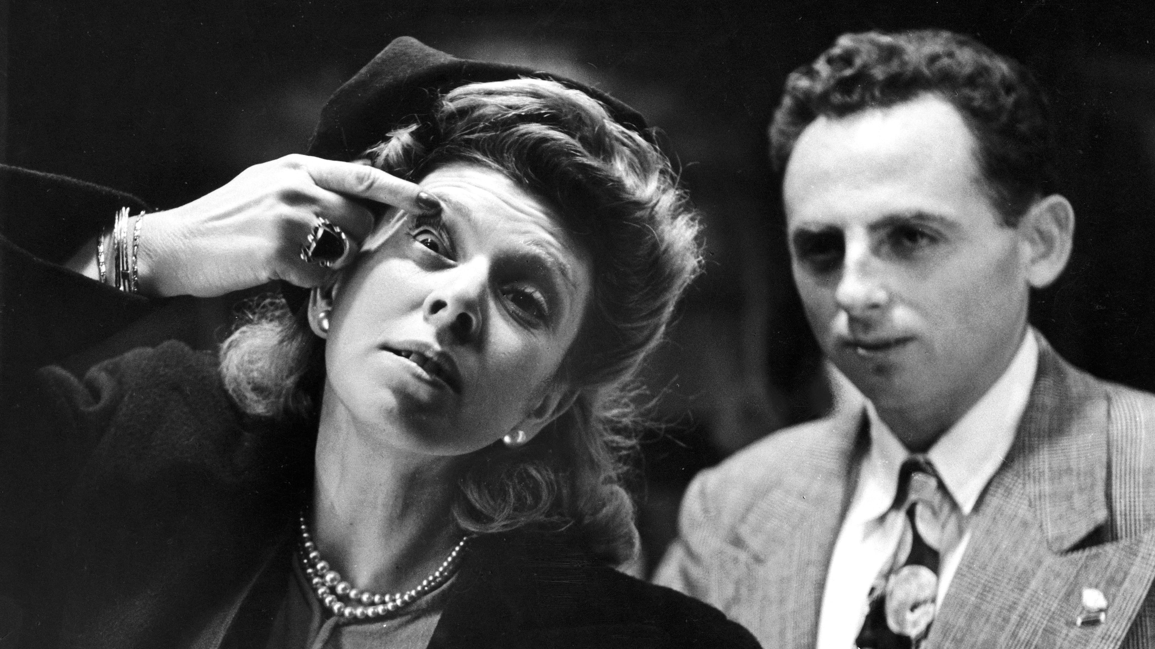 Old black and white photo of a woman checking if she has something in her eye in a mirror, photographed from the back of the mirror.